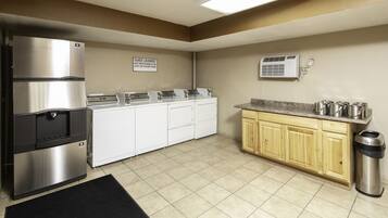 A kitchen with a stainless steel refrigerator, a water dispenser, a sink, a countertop with pots, a trash can, and a sign indicating guest laundry.