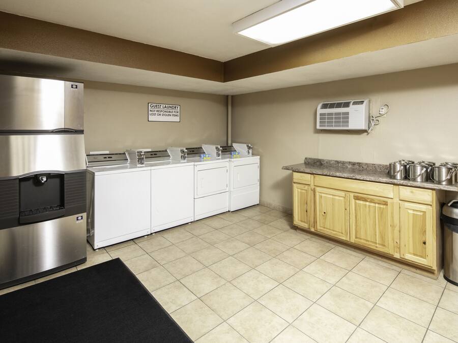 A kitchen with a stainless steel refrigerator, a water dispenser, a sink, a countertop with pots, a trash can, and a sign indicating guest laundry.