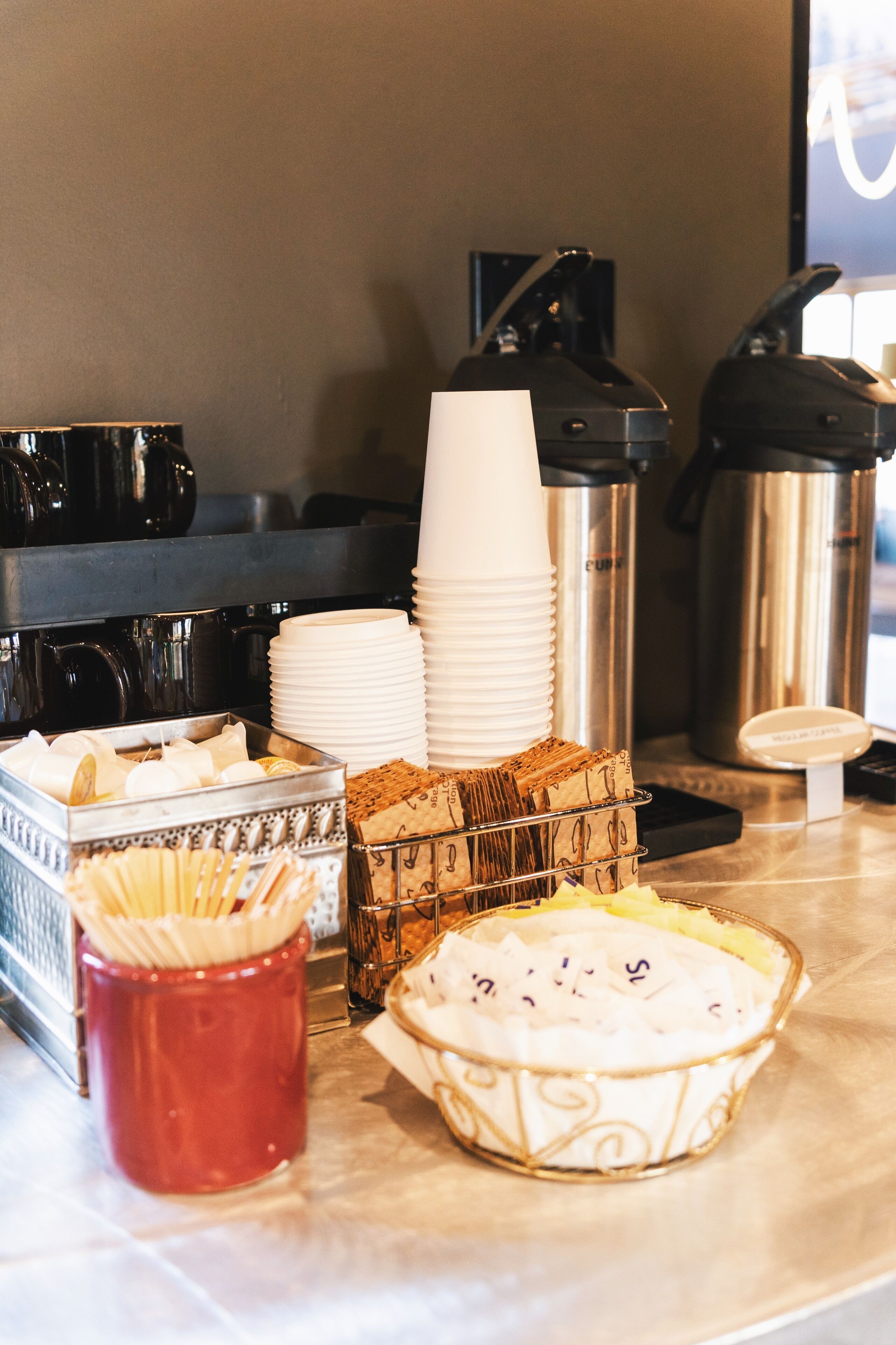 A coffee station with stacks of paper cups, a coffee machine, a thermos, and a bowl of cream with a spoon.