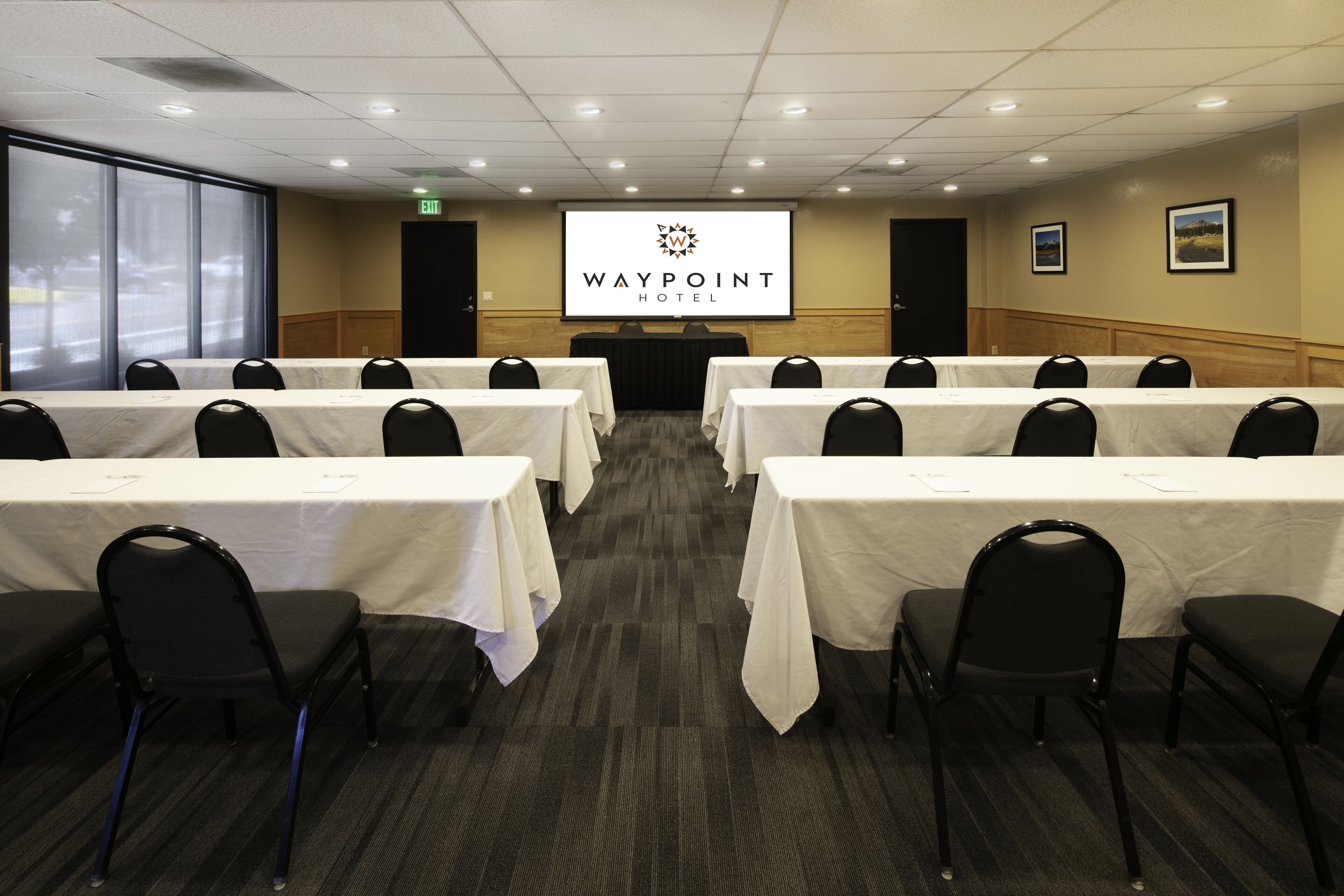 Een vergaderzaal met meerdere tafels en stoelen, een projectiescherm waarop het logo van Waypoint Hotel staat afgebeeld, en ingelijste foto's aan de muur.