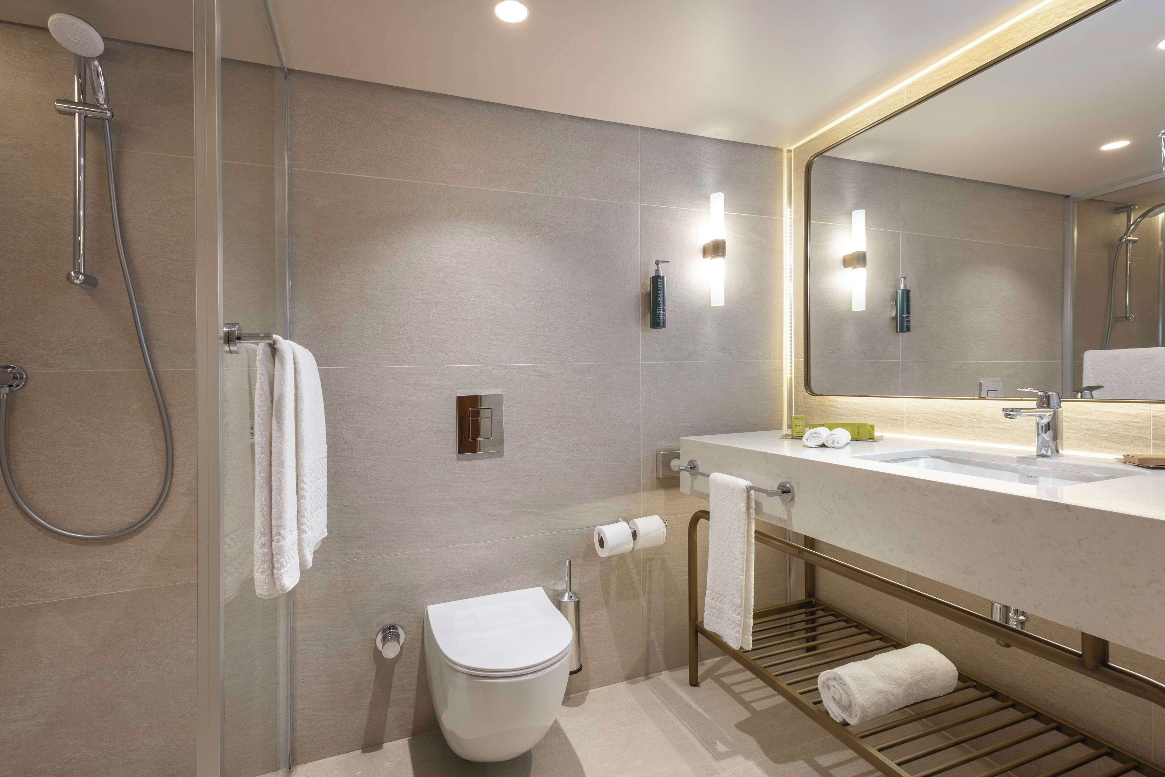 A modern bathroom with a glass-enclosed shower, a toilet, a sink with a towel rack, and a mirror with integrated lighting.