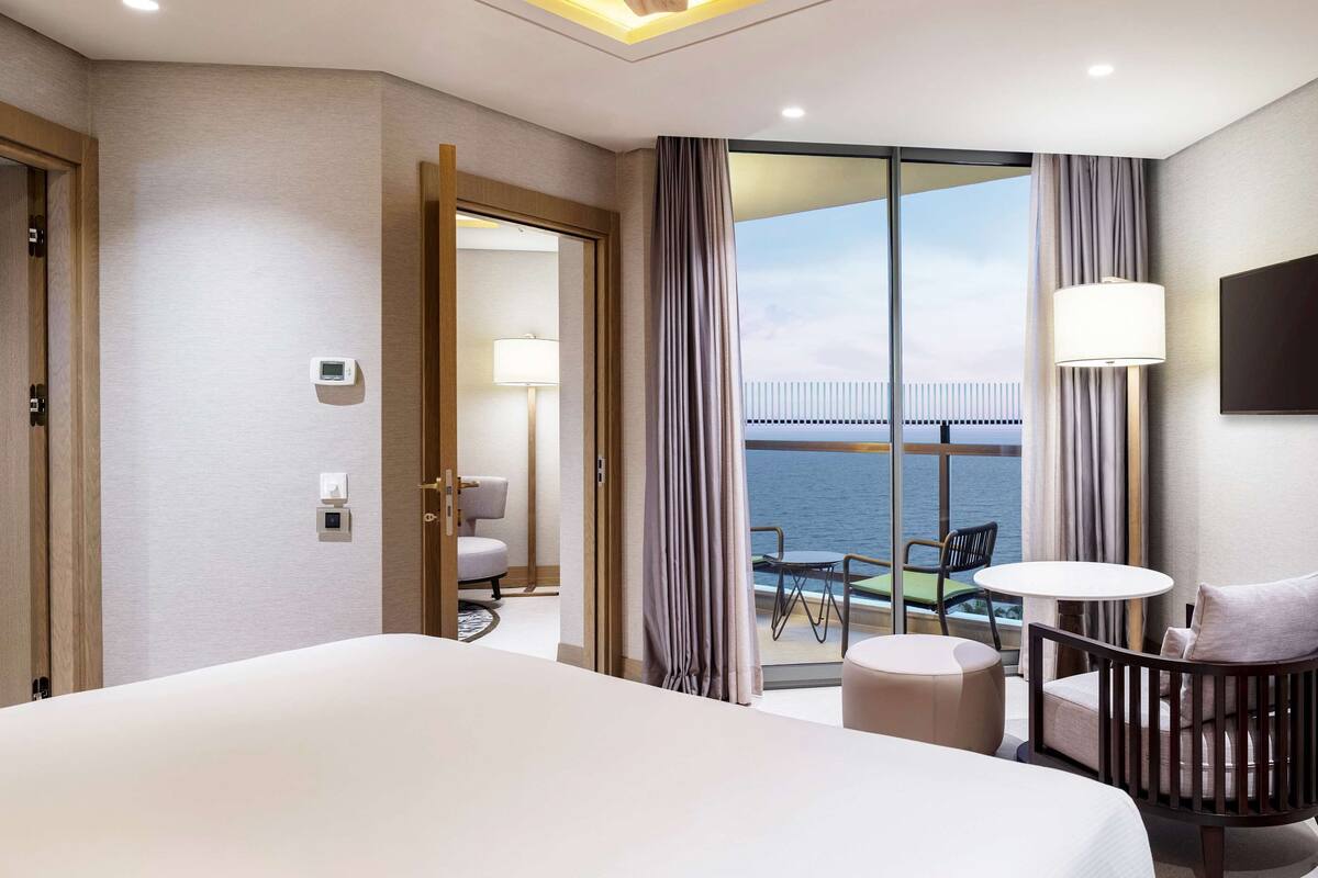 Quarto de hotel com uma cama grande, varanda com vista para o mar, televisão fixada na parede e uma pequena mesa com cadeiras.