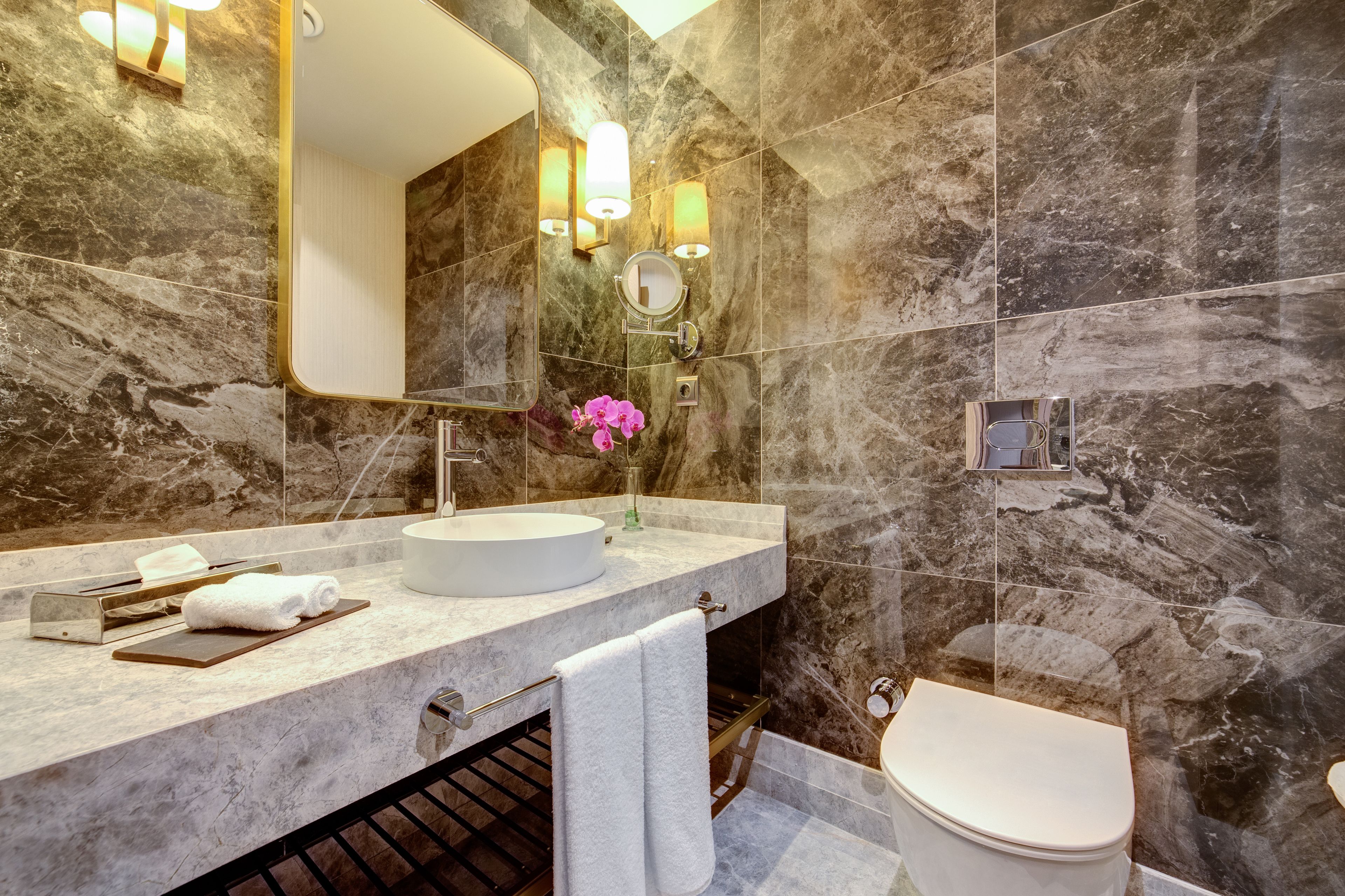 A modern bathroom with a marble wall, a round mirror, a white sink, and a toilet.