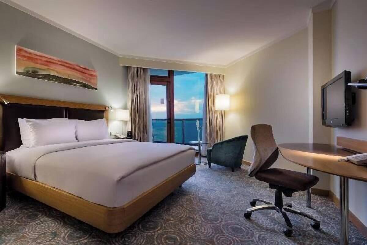 Quarto de hotel com cama, escrivaninha, cadeira, televisão e uma janela grande com cortinas.