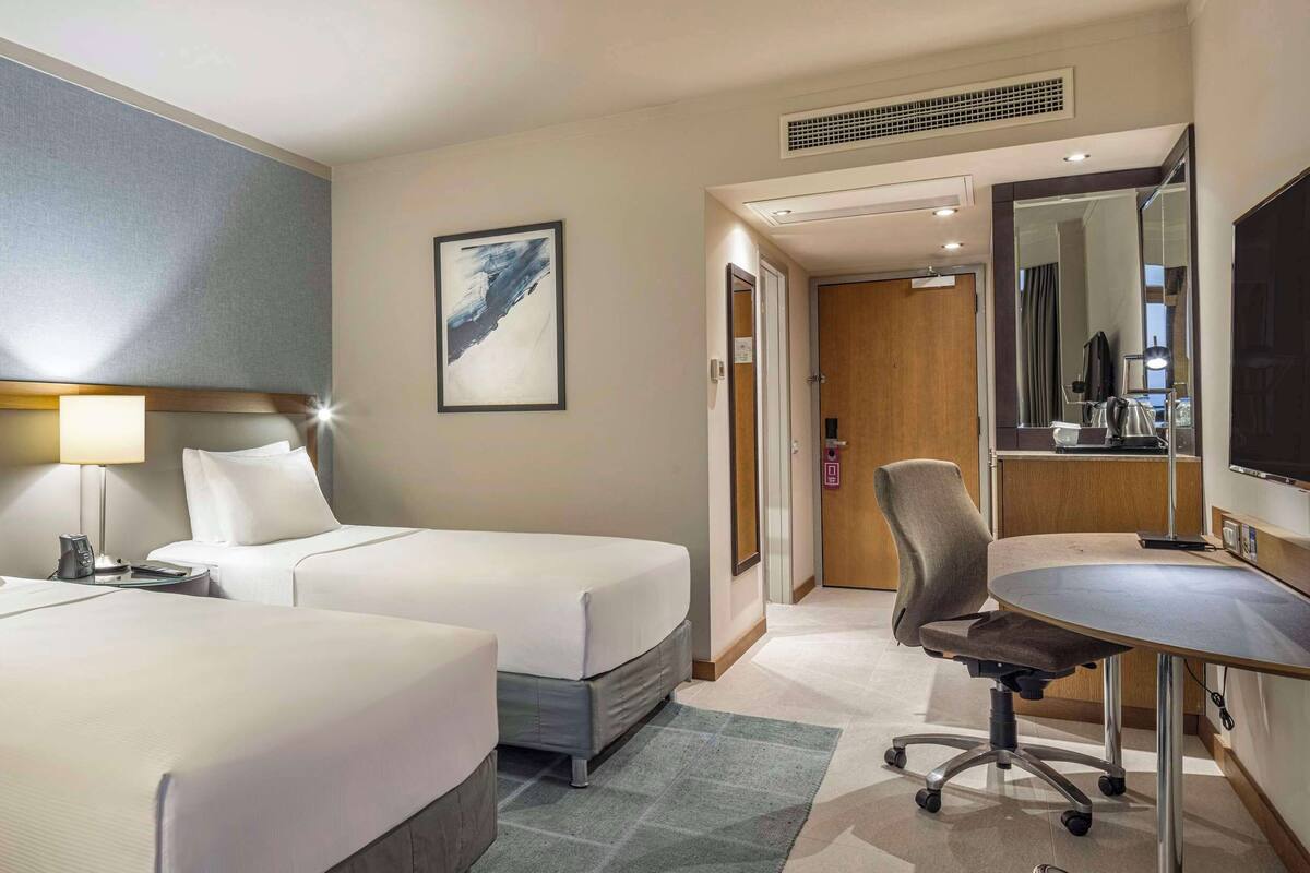 Quarto de hotel com duas camas, uma mesa de trabalho com cadeira, televisão e porta de banheiro.