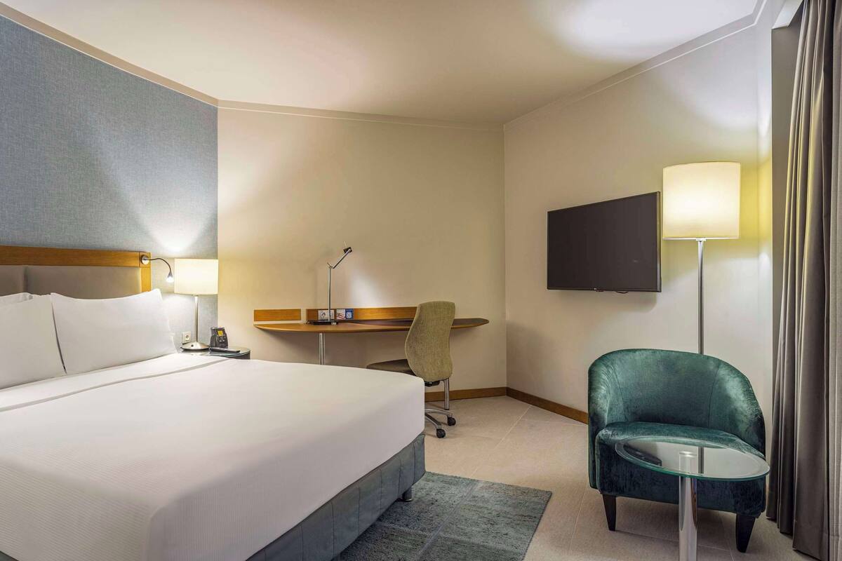 Quarto de hotel com cama, escrivaninha, cadeira, televisão e cortinas.