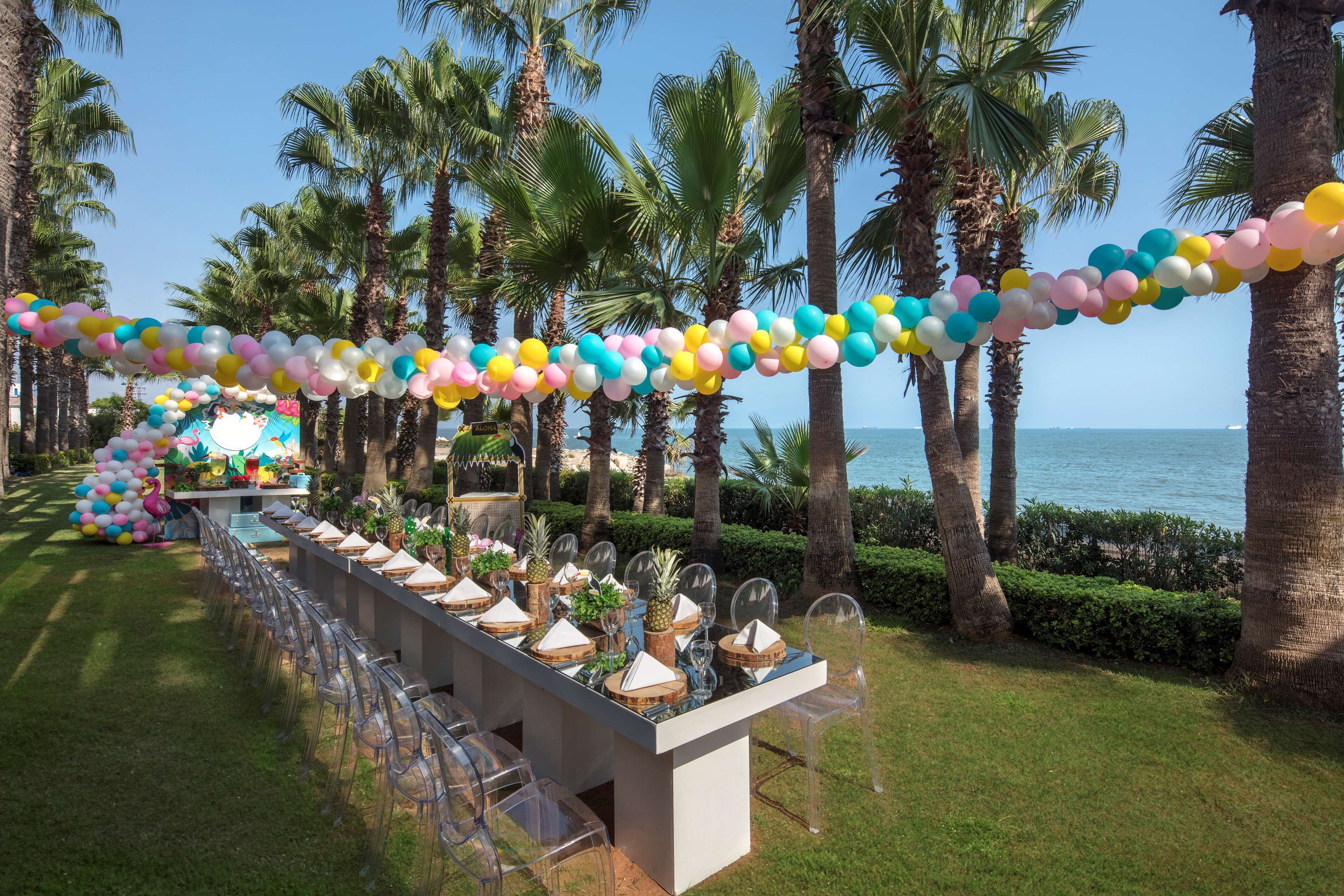 A long table set up outdoors with clear chairs, a decorative archway, and a view of the ocean.