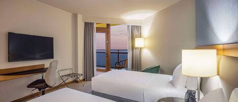 Une chambre d’hôtel avec un lit, une chaise, une télévision, une lampe et une vue sur la mer, visible à travers une grande fenêtre.