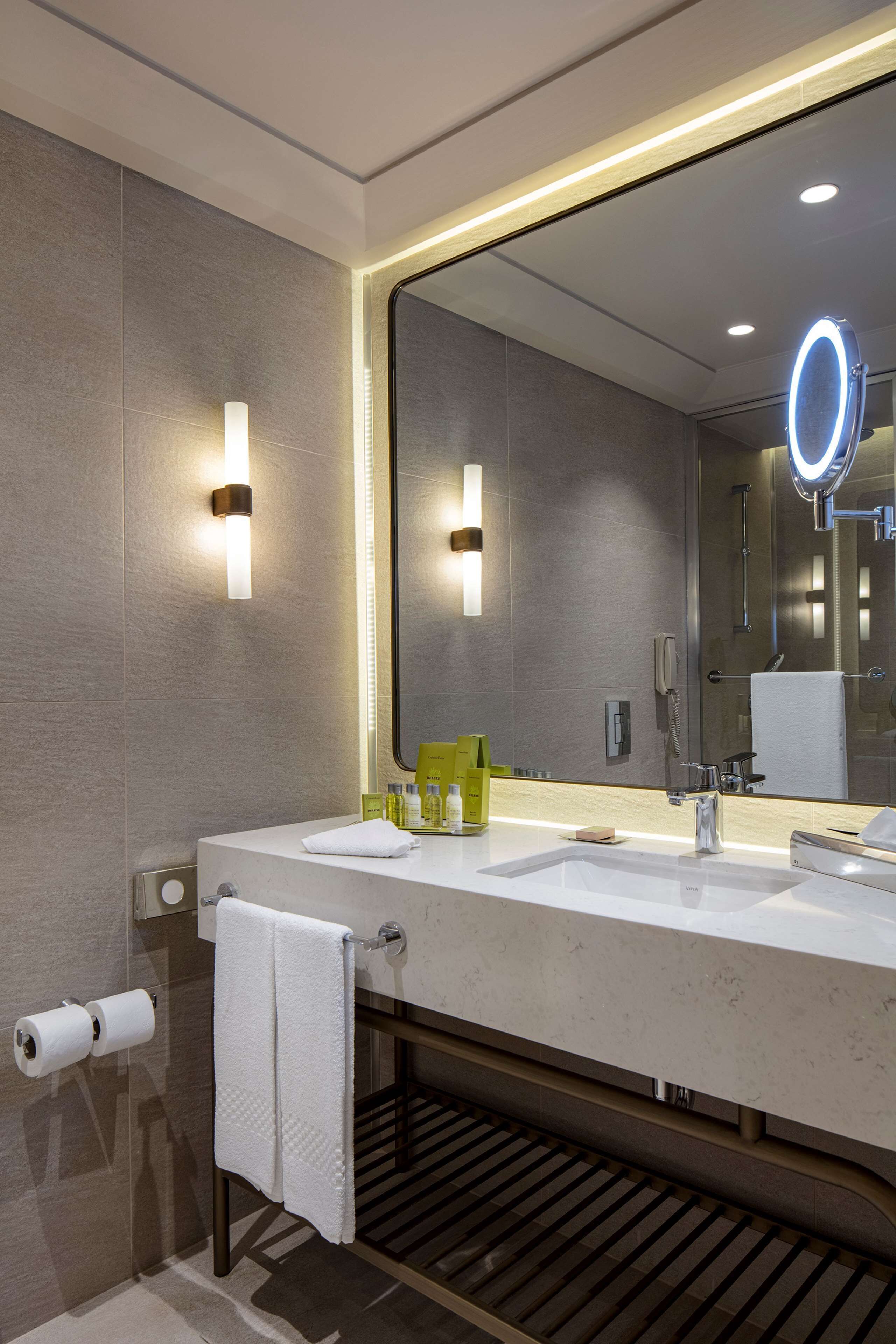 A modern bathroom with two sinks, a large mirror with a built-in light, and a toilet paper holder.