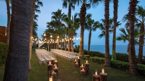A wedding ceremony setup with chairs arranged in rows, string lights, and candles on wooden stumps, set against a backdrop of palm trees and a view of the ocean. - Mersin HiltonSA (Mersin)