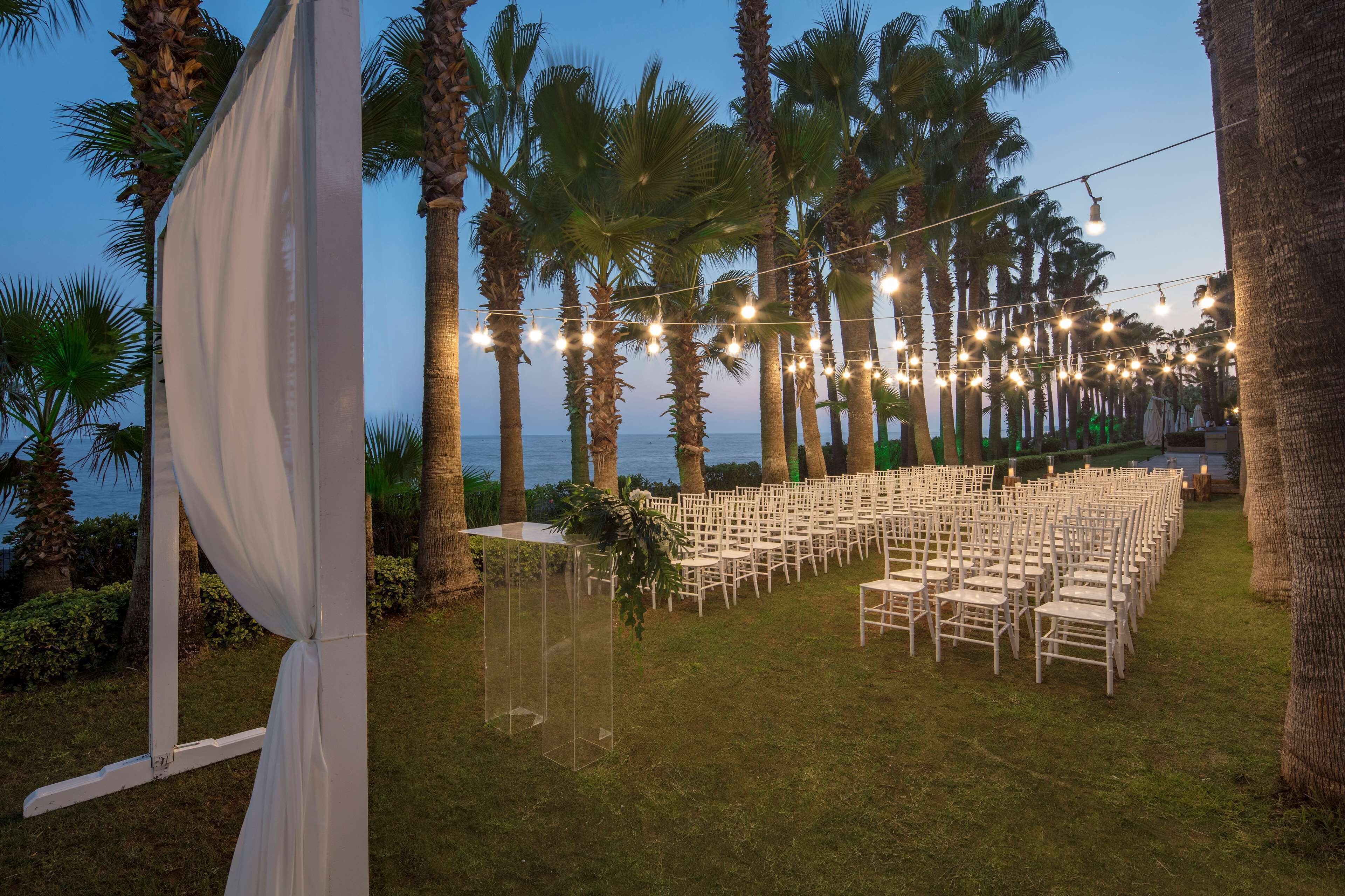 An outdoor wedding venue with rows of white chairs and tables, palm trees, and string lights.