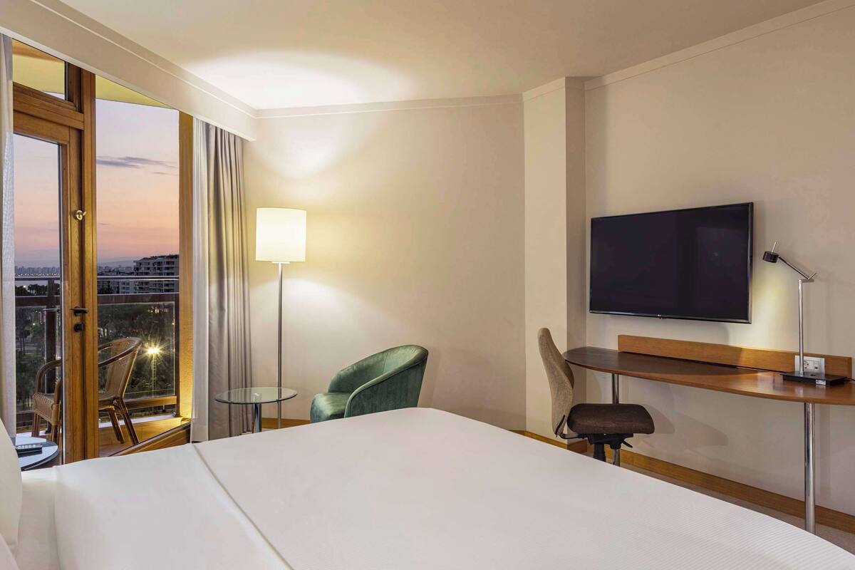 Quarto de hotel com cama, escrivaninha, televisão, poltrona e vista da cidade ao pôr do sol.