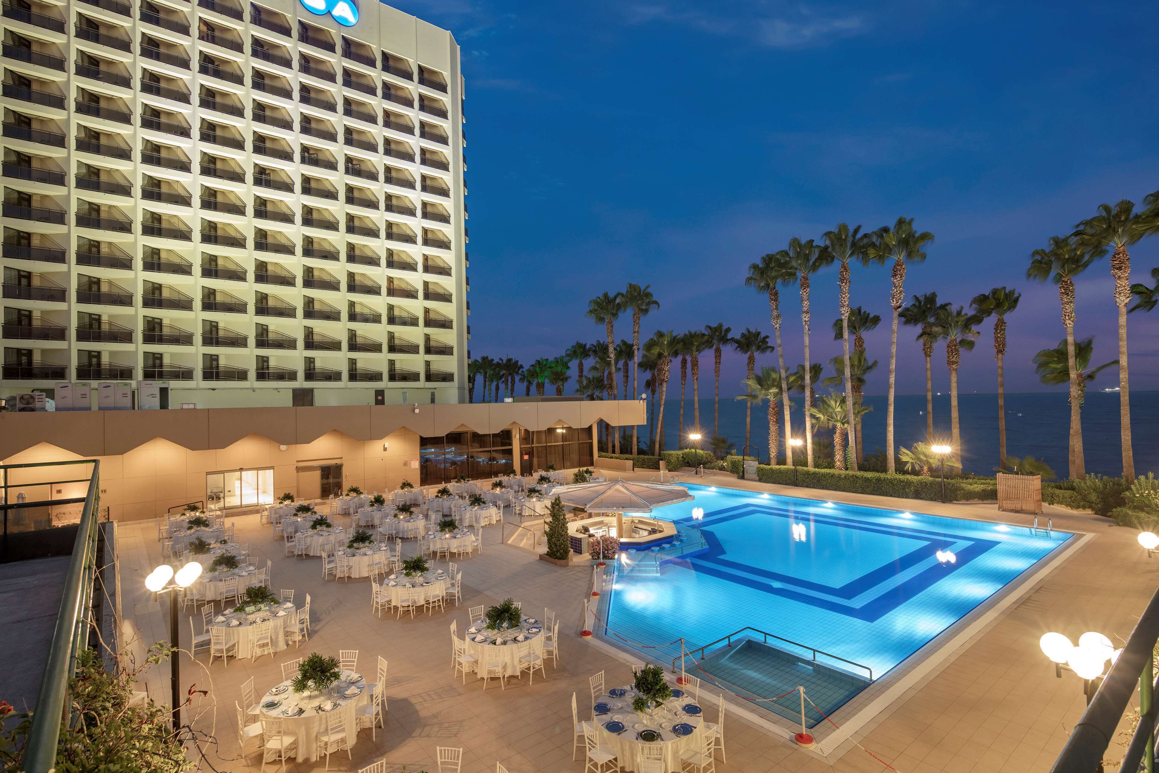 A hotel pool area with multiple tables and chairs, a large building in the background, and palm trees.