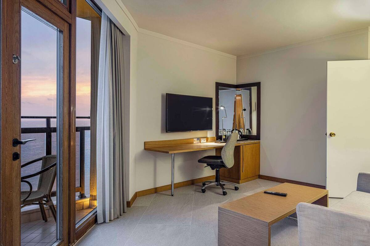 Quarto de hotel com varanda, escrivaninha, cadeira, sofá, televisão e luminária.