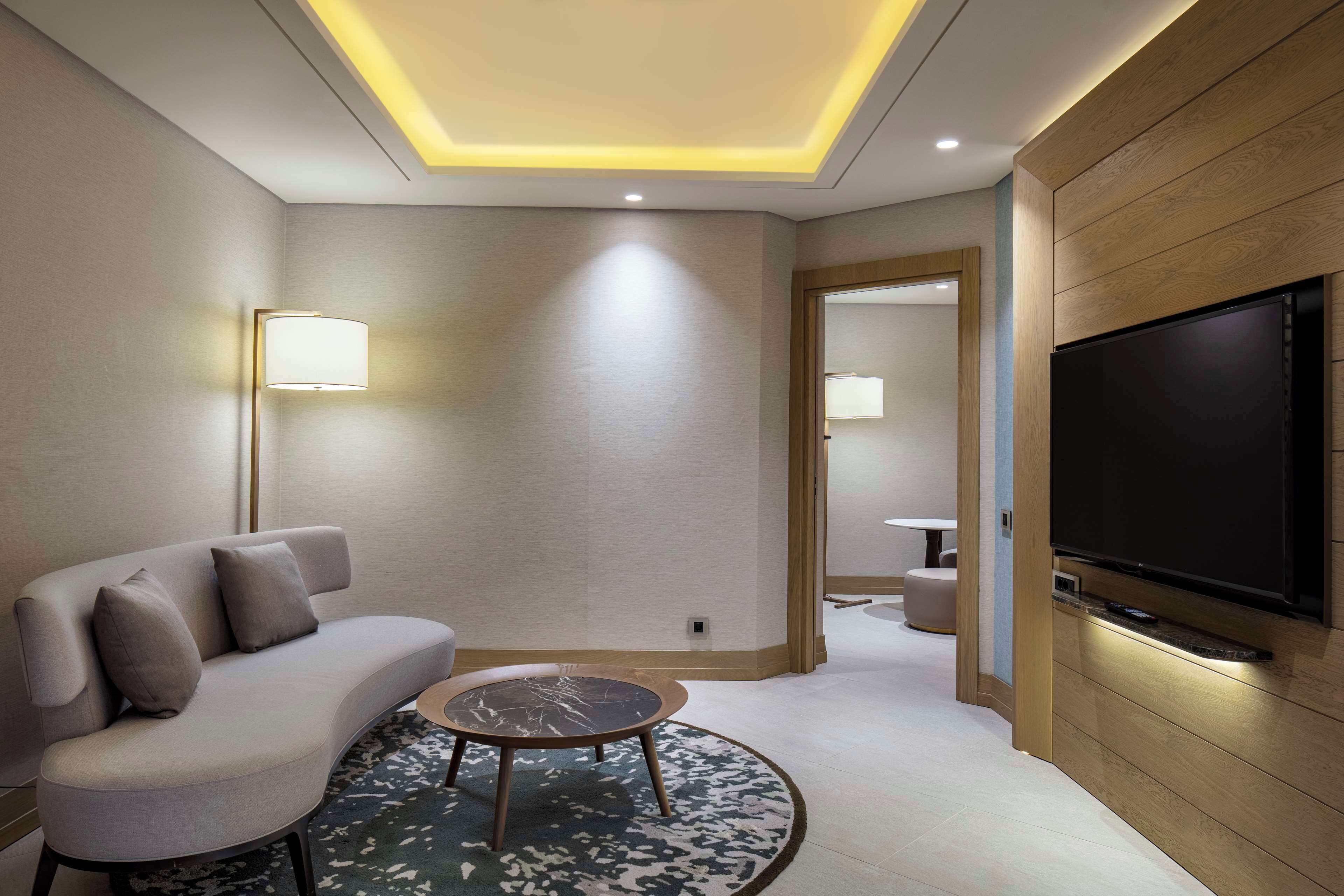 A modern hotel room with a sofa, a round coffee table, a flat-screen TV mounted on the wall, and a ceiling light fixture.