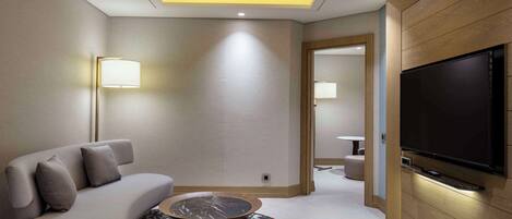 A modern hotel room with a sofa, a round coffee table, a flat-screen TV mounted on the wall, and a ceiling light fixture.