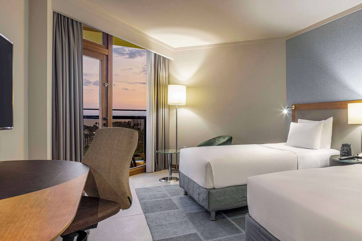 Quarto de hotel com cama, escrivaninha, cadeira, televisão e vista para a paisagem urbana.