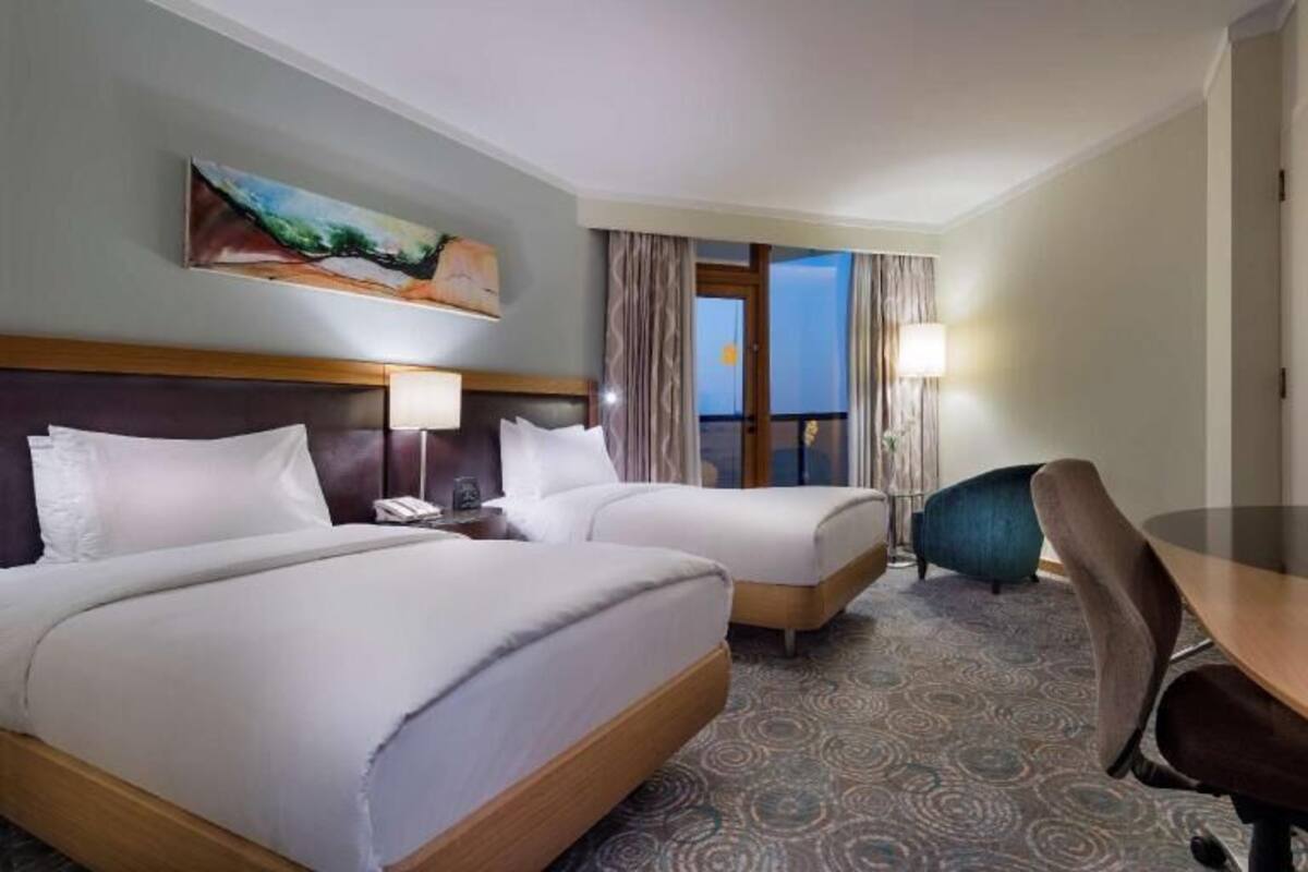 Quarto de hotel com duas camas, uma escrivaninha, uma televisão e um quadro na parede.