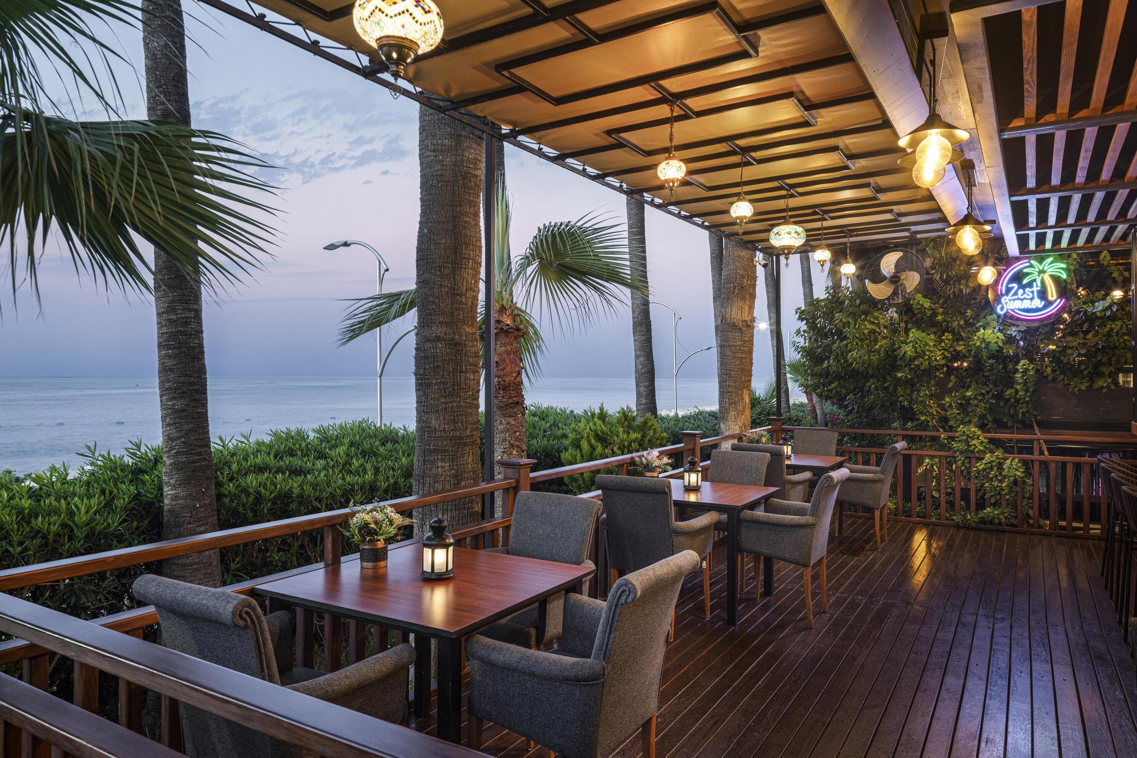 An outdoor dining area with wooden tables and chairs, surrounded by palm trees and a view of the ocean.