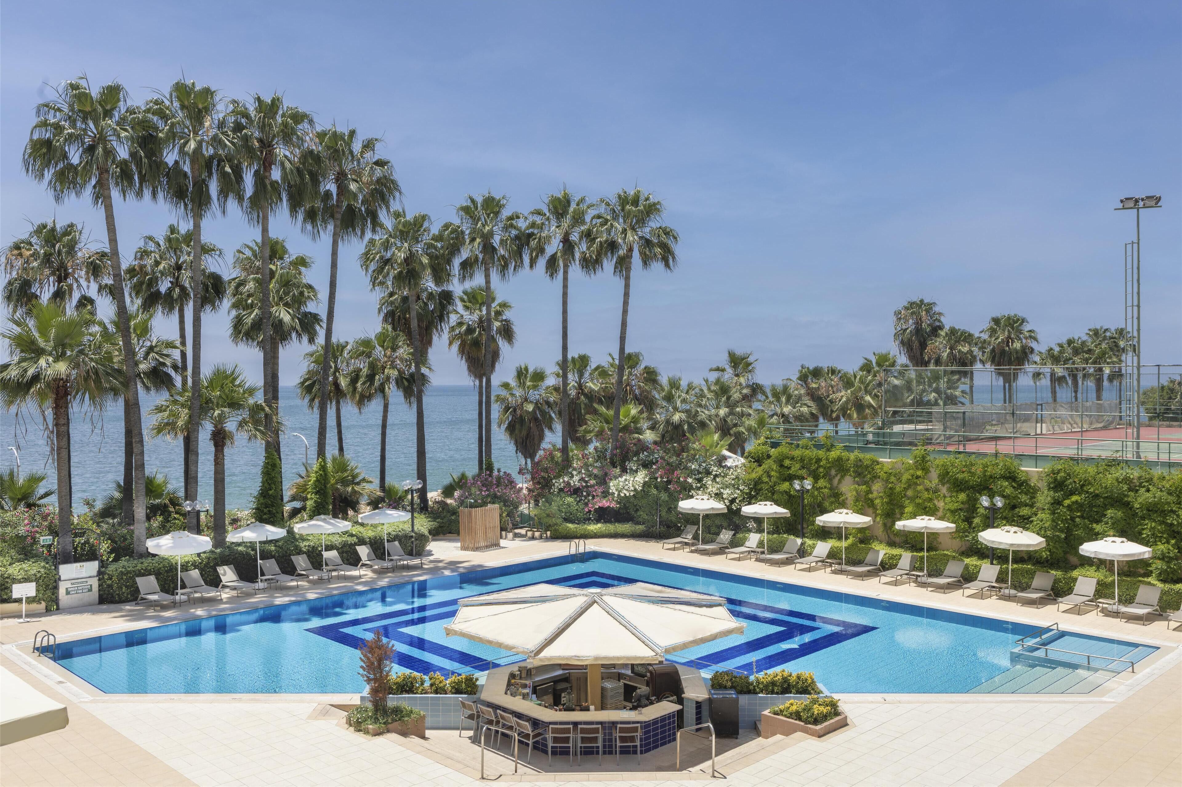 A pool area with lounge chairs and umbrellas, surrounded by palm trees and a view of the ocean.