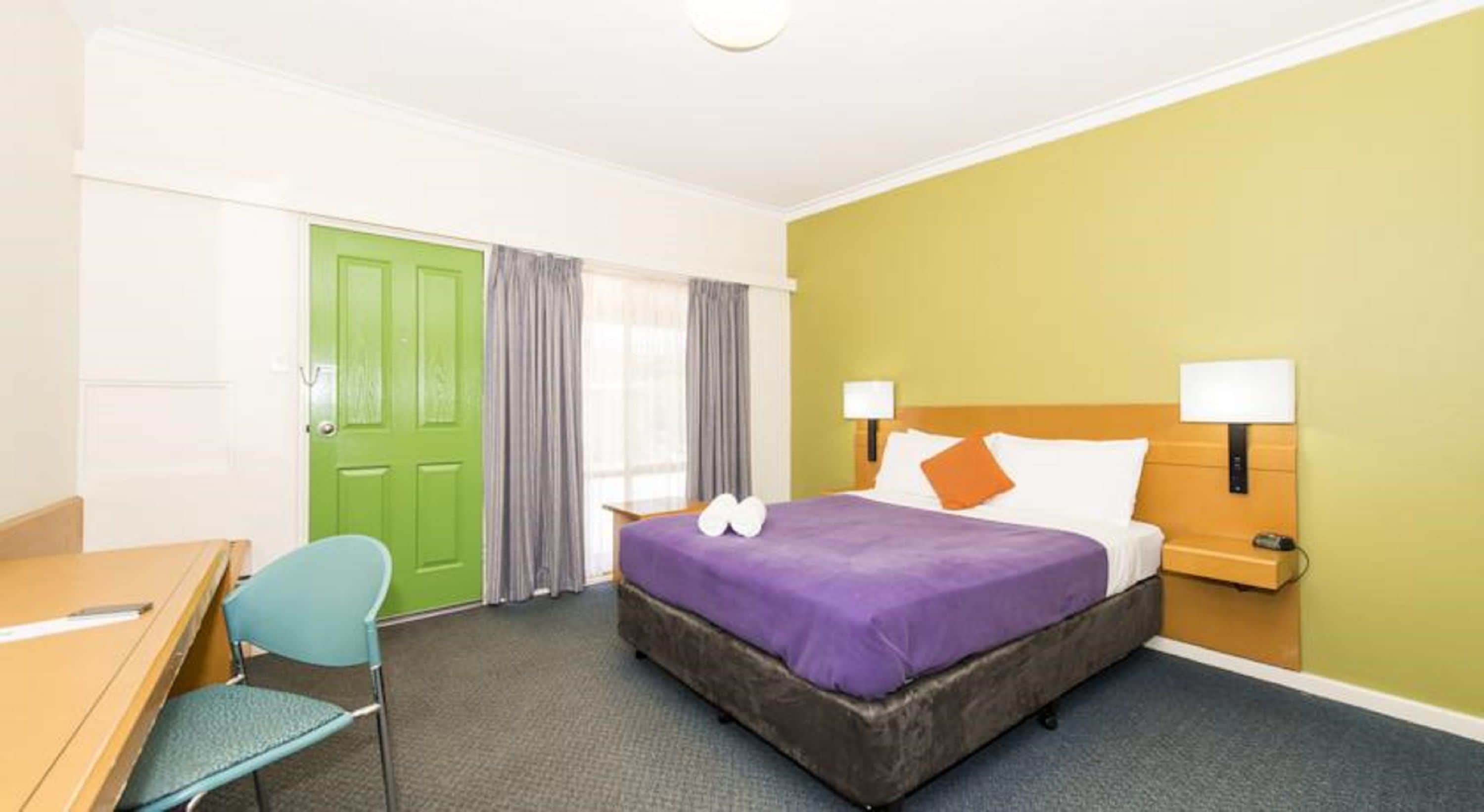 A hotel room with a bed, a desk, a chair, and a green door.