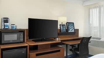 Habitación de hotel con escritorio de madera, televisión de pantalla plana, microondas, cafetera, refrigerador, una silla, una lámpara y una ventana con cortinas.