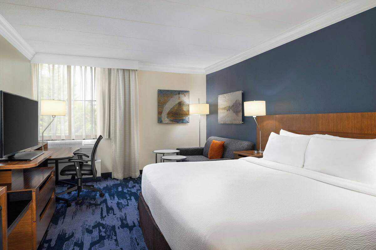 Habitación de hotel con una cama grande, un escritorio con computadora, una silla, una mesita y una lámpara.