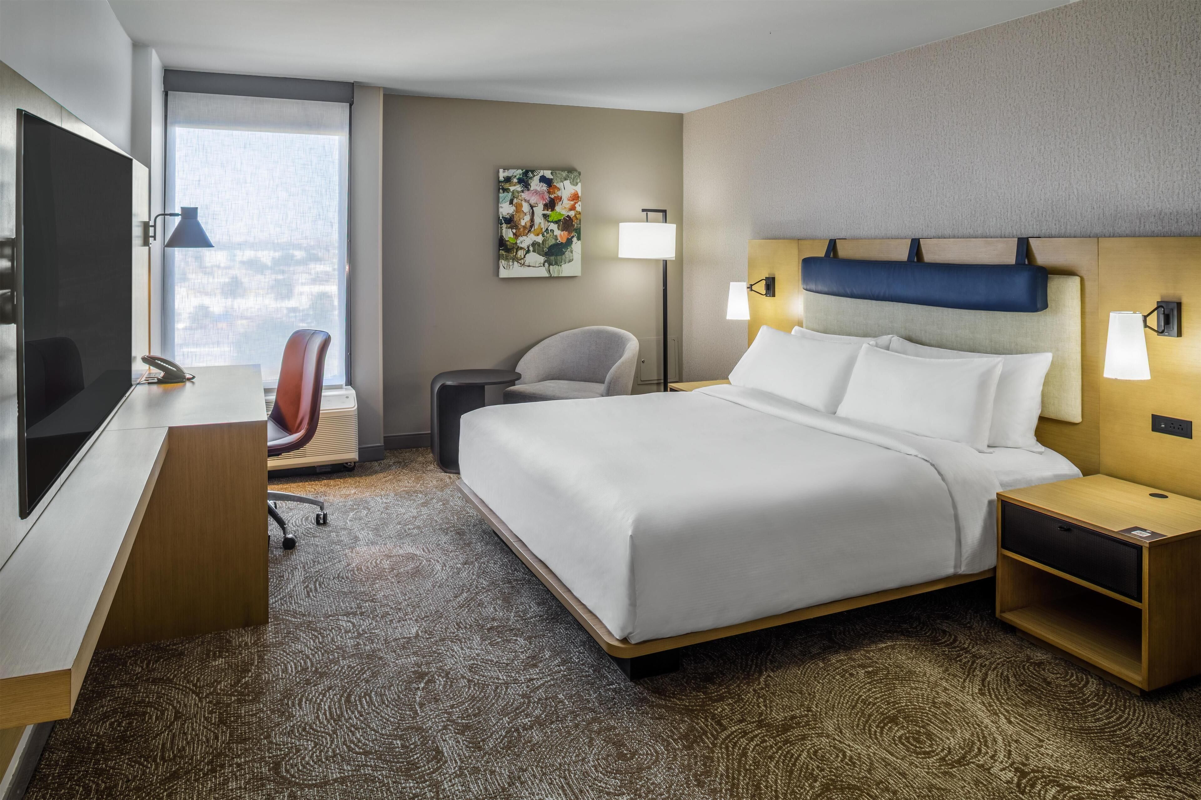 A hotel room with a bed, desk, chair, and a large window.