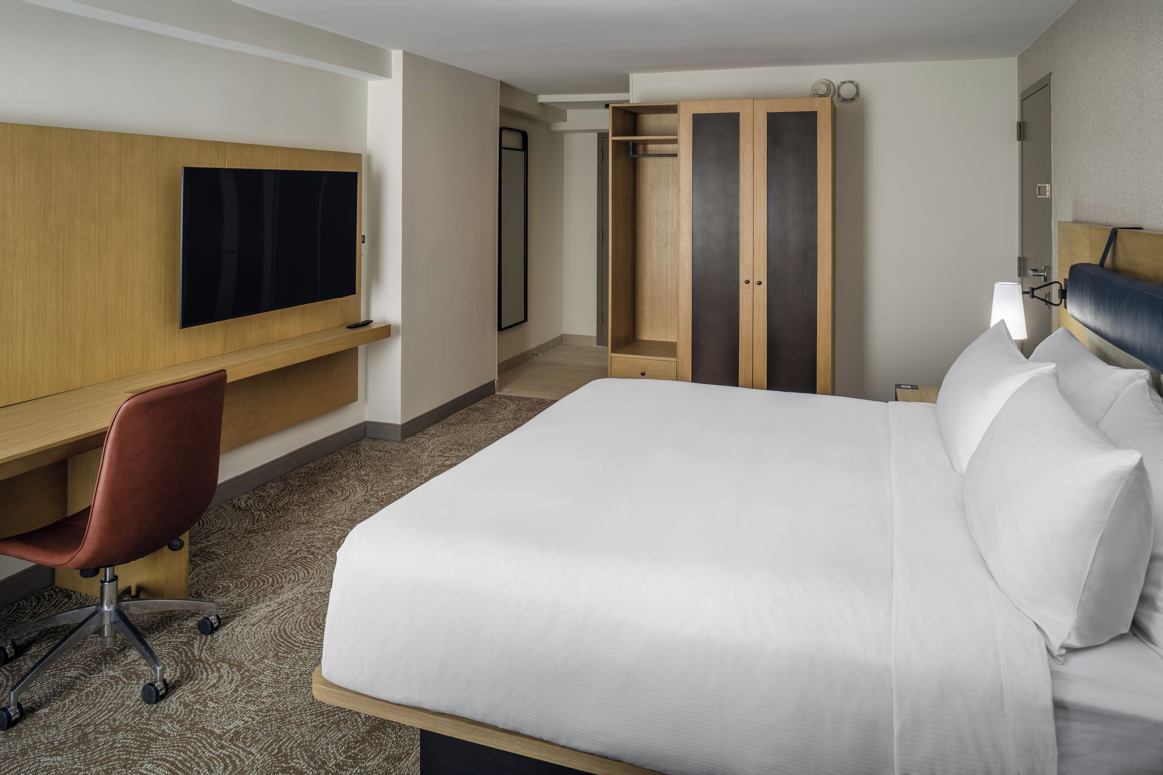 A hotel room with a large bed, a desk with a chair, a TV, a wardrobe, and a bedside table with lamps.