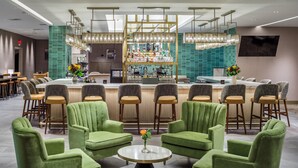 A modern bar area with a bar counter, green upholstered chairs, and a round table with a vase of flowers. - DoubleTree by Hilton Laredo (Laredo)