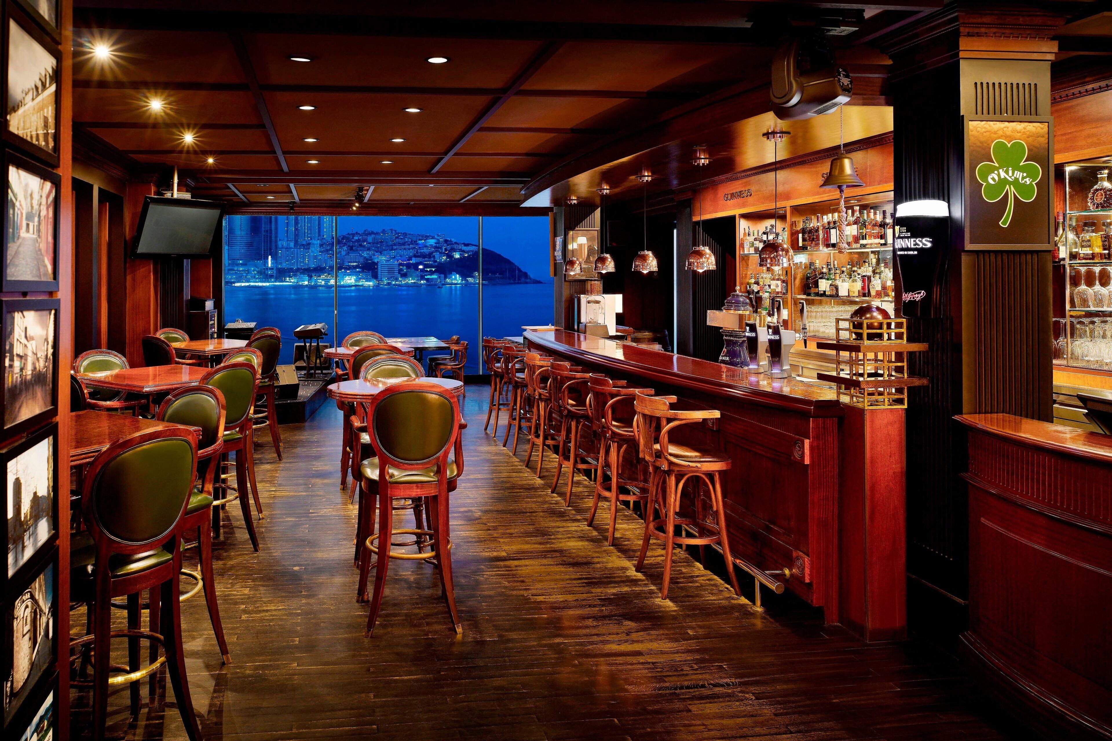 A bar area with wooden furniture, a large screen, and a view of a cityscape.