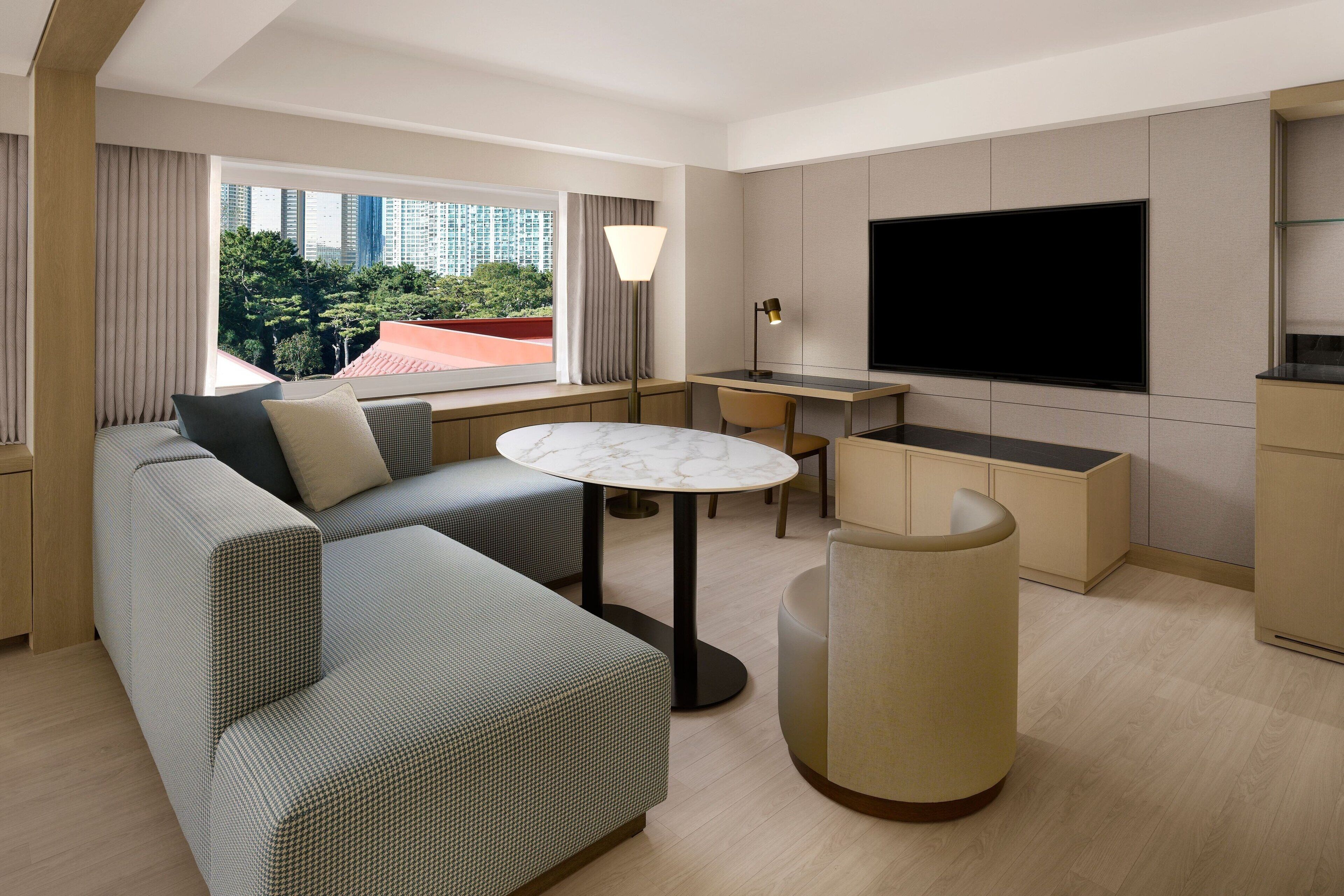 A modern hotel room with a sofa, a round table, a TV, and a view of the cityscape.