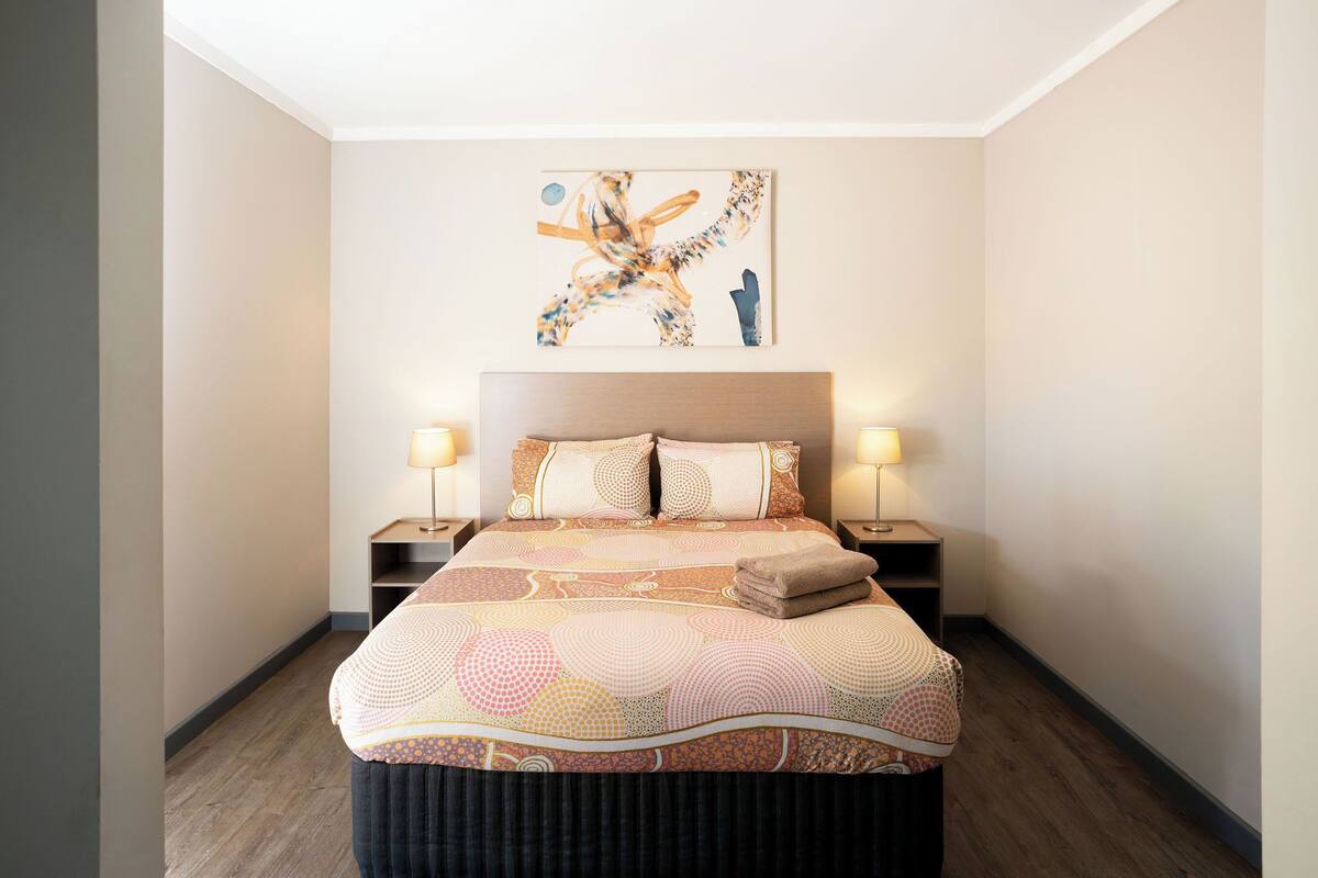 A neatly made bed with a patterned bedspread, two bedside lamps, and a wall-mounted abstract painting.