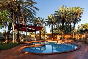 Una piscina con toldo rojo, rodeada de palmeras y un patio con mobiliario exterior.