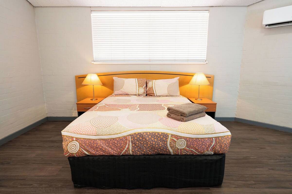 A bedroom with a bed, two bedside tables, a window, and an air conditioning unit.