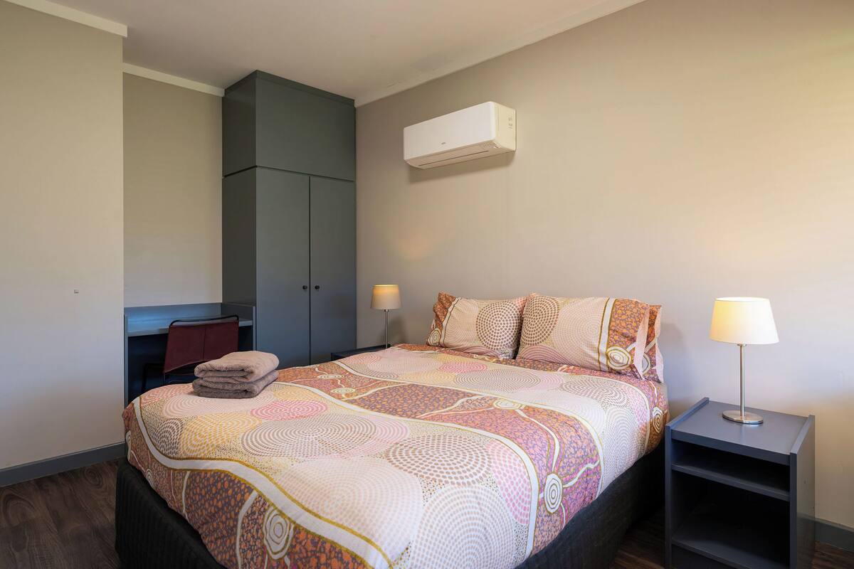 A bedroom with a bed, a desk, a wardrobe, and an air conditioner.