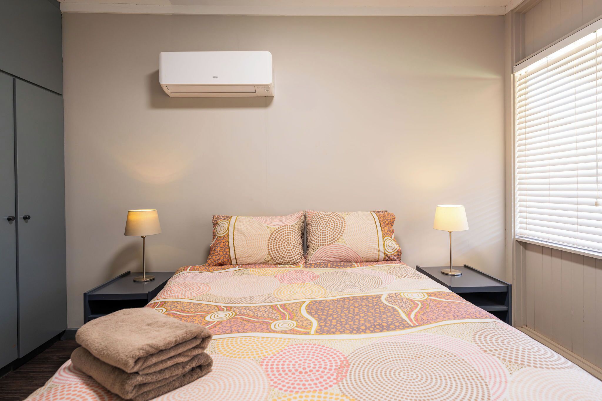 Una cama bien hecha con ropa de cama estampada, un aire acondicionado de pared, lámparas de noche y una ventana con persianas.