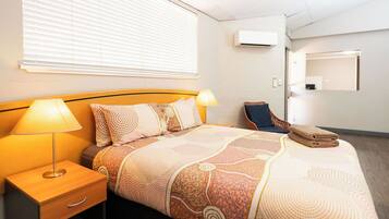 A hotel room with a bed, two bedside lamps, a chair, a folded towel, and an air conditioning unit on the wall.