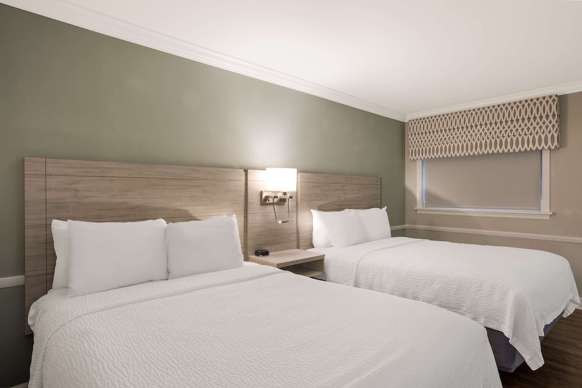 Habitación de hotel con dos camas, una mesita de noche, una lámpara y una ventana con cortinero.