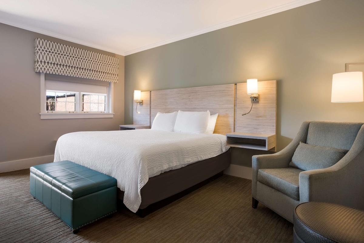 A hotel room with a large bed, a green ottoman, a grey armchair, and two bedside tables with lamps.