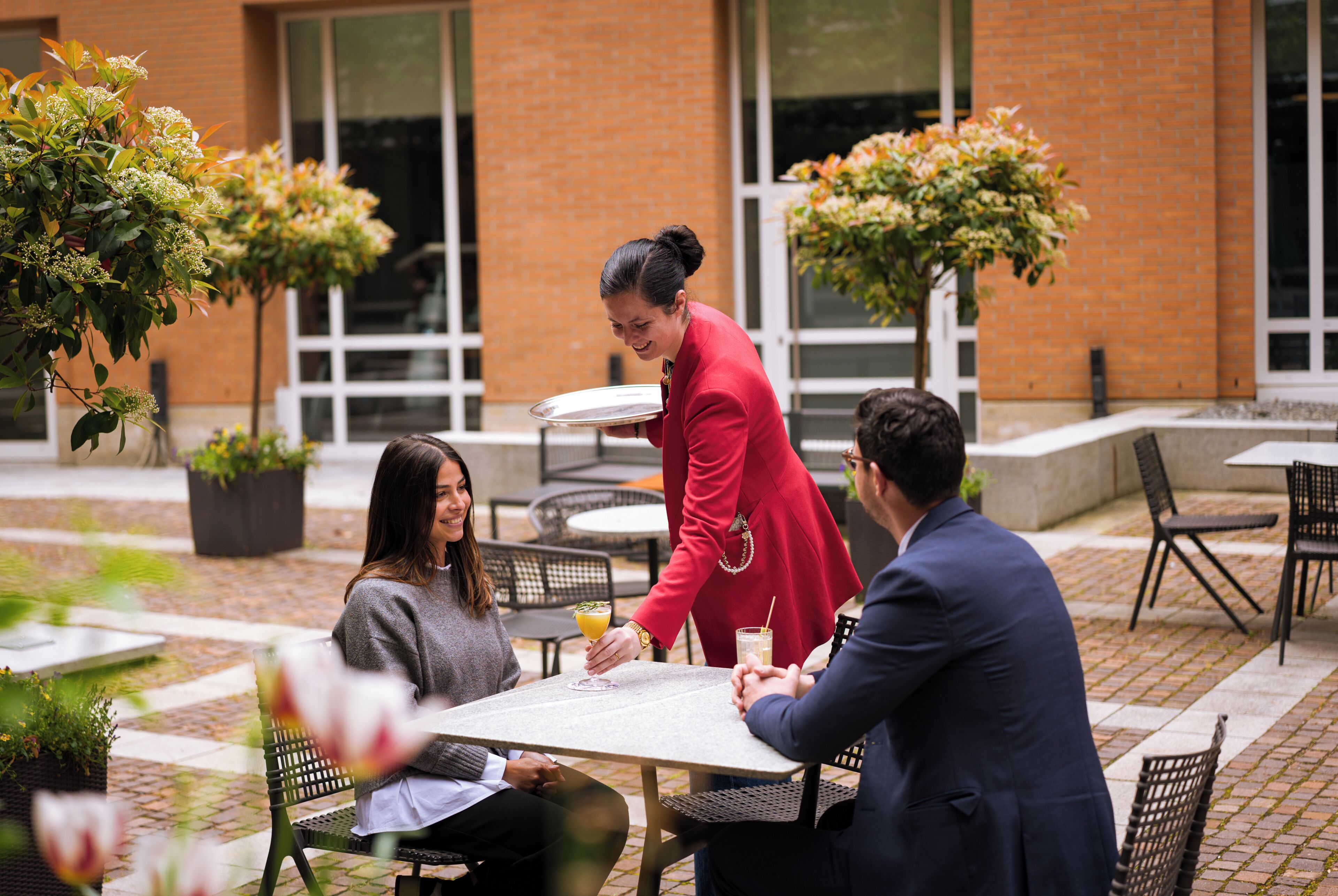 An outdoor seating area with tables and chairs, a waiter serving drinks, and potted plants.
