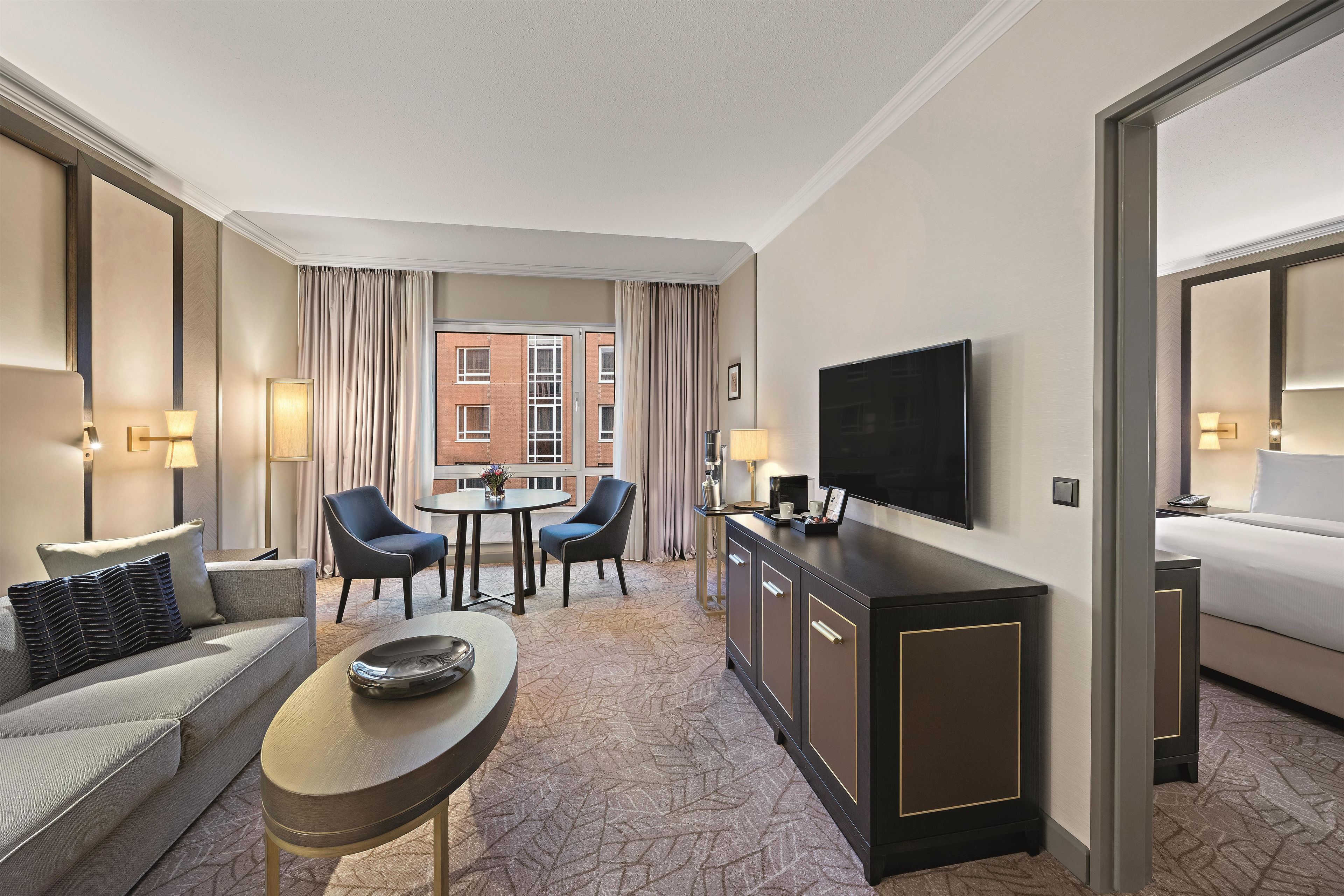 Habitación de hotel con cama, sofá, mesa de comedor con sillas, televisión y vista a un edificio por la ventana.