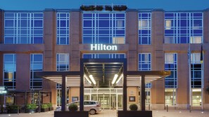 Entrada de un hotel Hilton con acceso cubierto, puertas de vidrio y un coche estacionado frente.