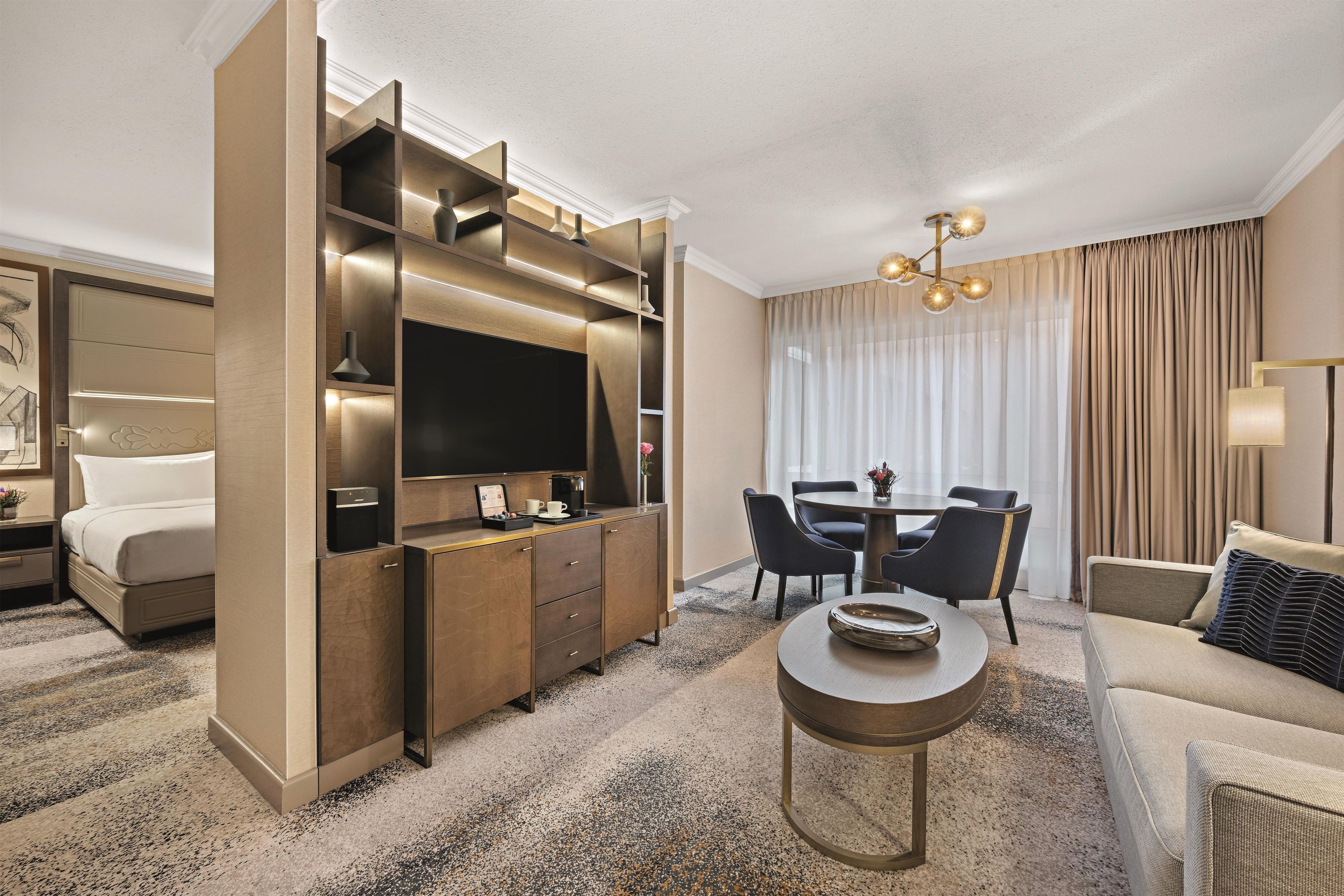A modern hotel room with a bed, a desk with a flat-screen TV, a dining area with chairs, and a sofa.