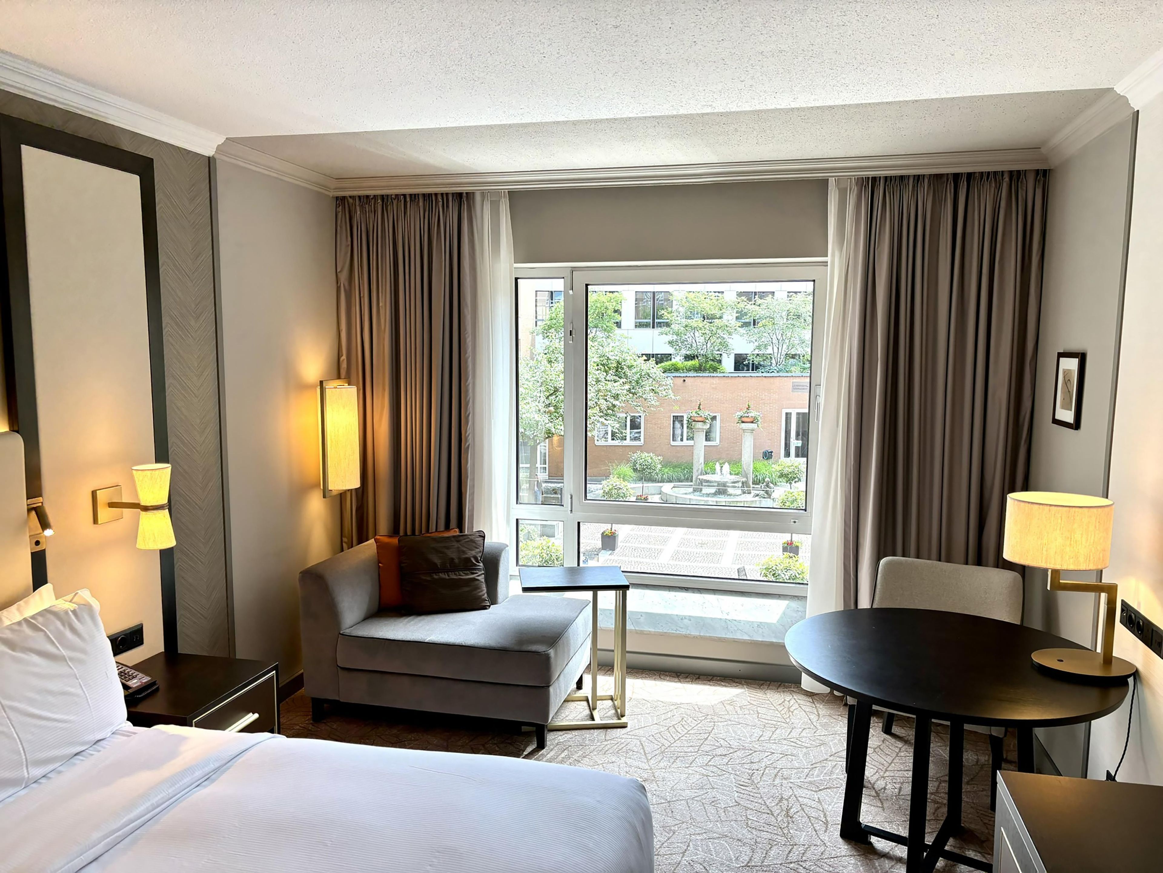 A hotel room with a bed, a sofa, a small table, and a view of the outside.