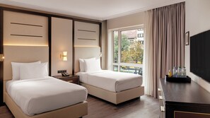 Habitación de hotel con dos camas, una mesita de noche con teléfono, una ventana con cortinas y un escritorio con televisor y botellas de agua.