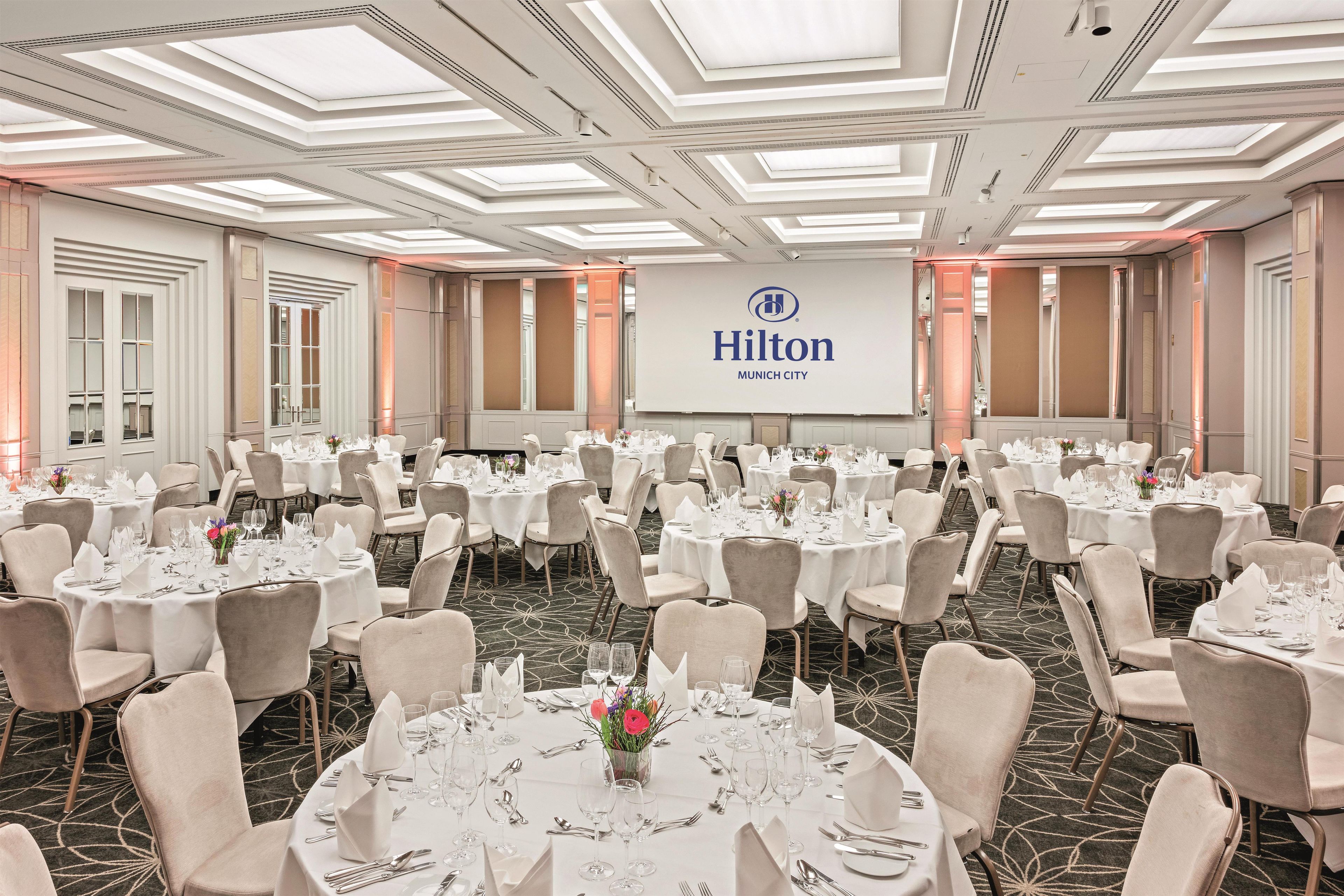 A large, well-lit conference room with multiple round tables set for dining, featuring a 'Hilton Munich City' sign on the wall.