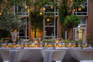 An outdoor dining area with a table set for a meal, surrounded by greenery and a building in the background. - Hilton Munich City (Munich)