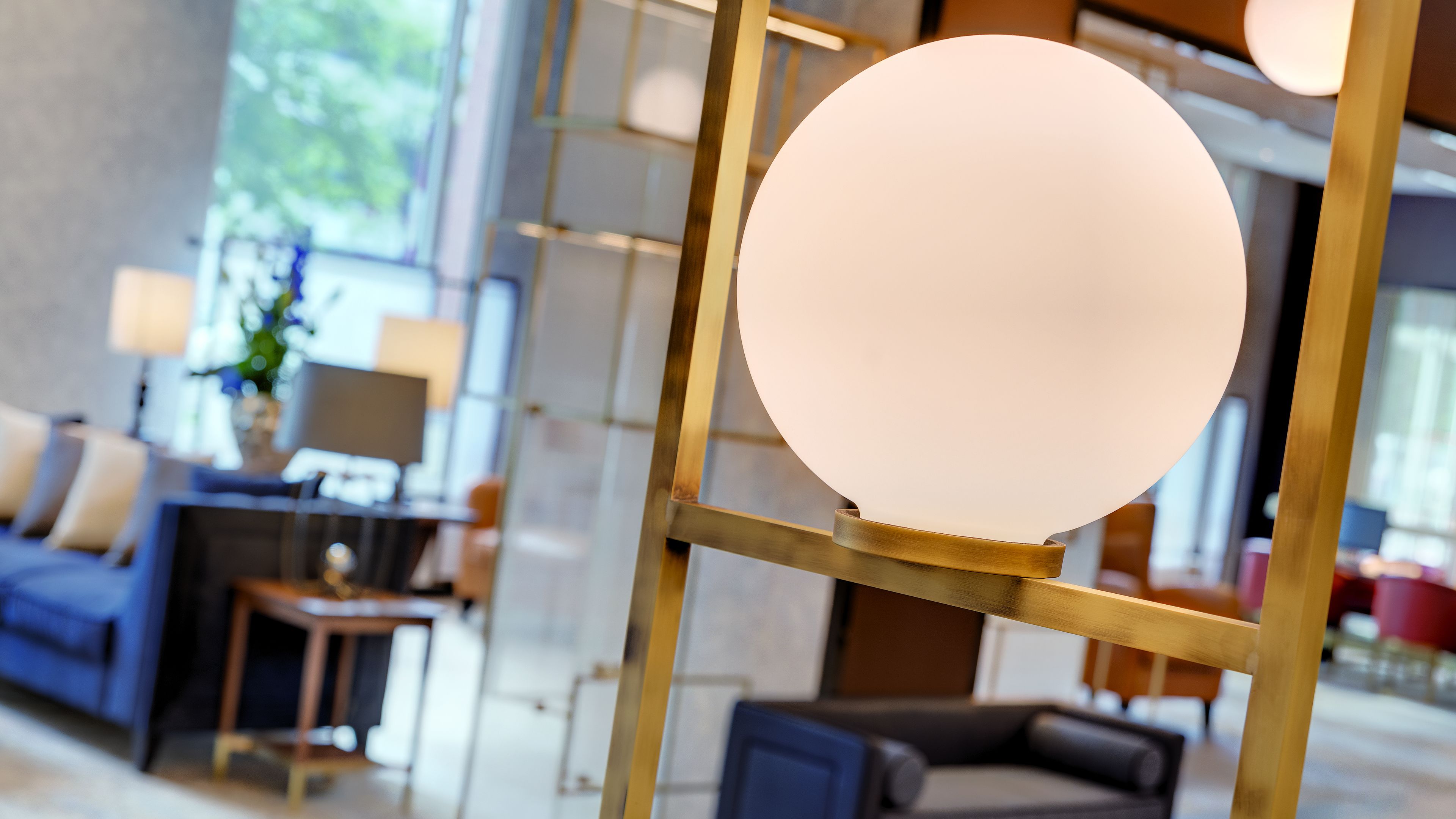 A modern interior with a round light fixture on a gold stand, a blue sofa, and a desk with a lamp.