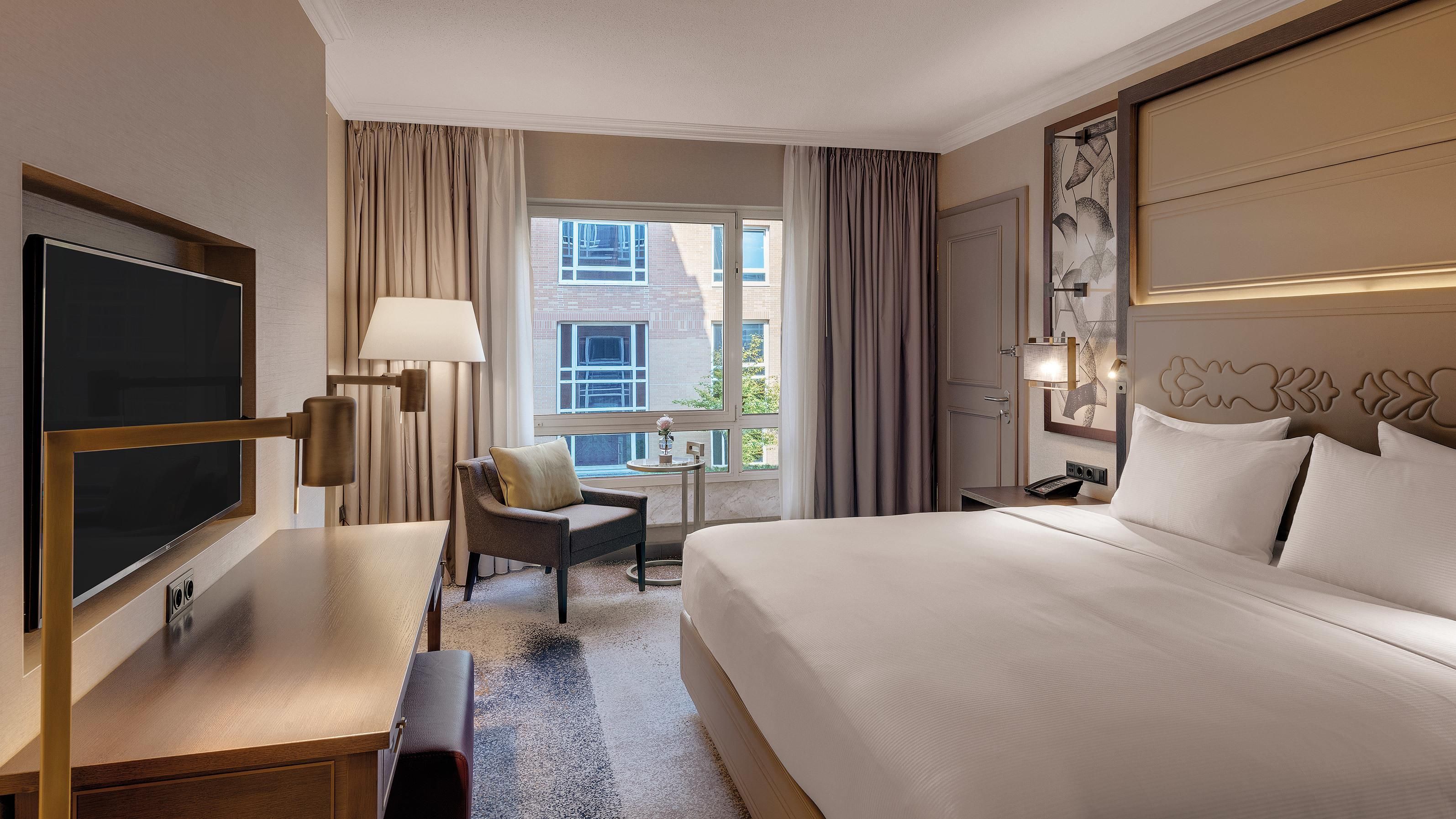 Habitación de hotel con cama, escritorio, silla, televisor, lámpara y ventana con cortinas.